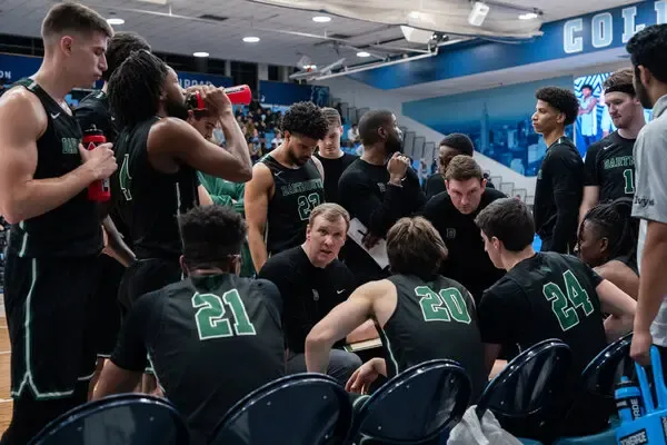 In the image, a diverse group of college student athletes is gathered on a college campus, discussing their experiences with athletic scholarships and financial aid in U.S. college athletics. They are engaged in conversation about the recruiting process and the various types of scholarships available, including full ride scholarships and partial scholarships, highlighting the importance of academic success alongside athletic ability.