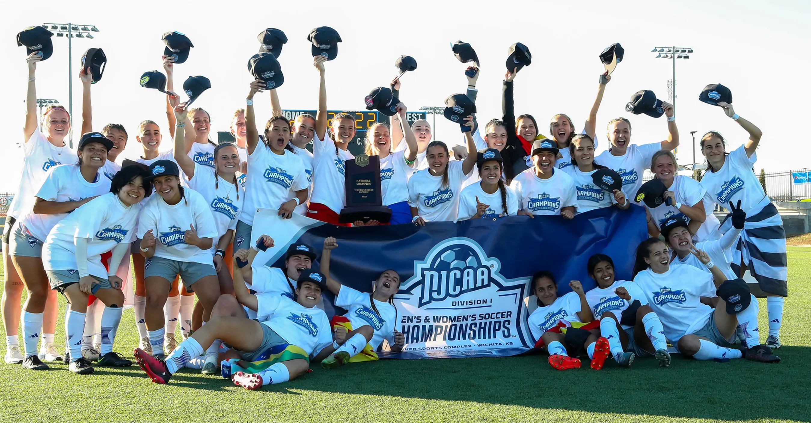 Women's team wearing white 'Champions NJCAA Division I' shirts and hats, clustered behind a blue championship banner